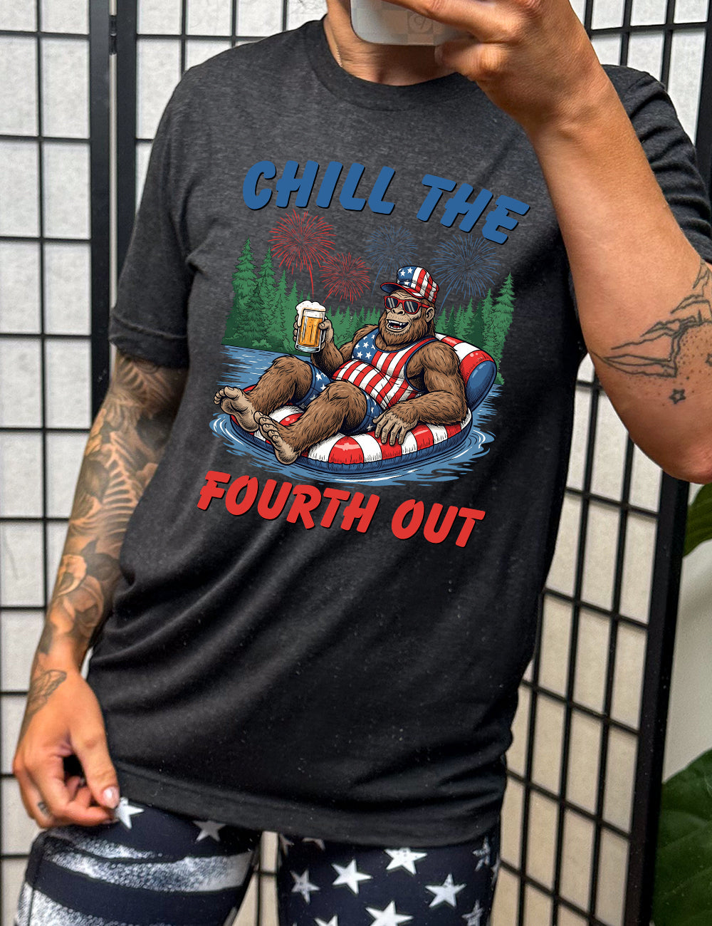 black unisex tee with the graphic of a grinning Bigfoot holding a beer, wearing sunglasses and an American flag outfit, in an American flag tube, in the water, with a background of trees, with fireworks above, and the words saying "chill the fourth out" on it