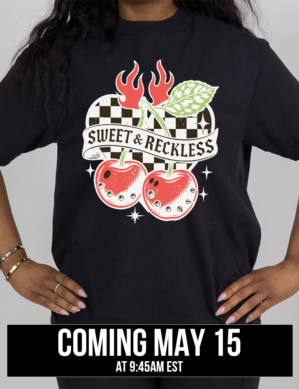 black modern tee with print placement on the front, with the graphic of two large cherries, with spikes, on connected stems with a leaf, in front of a checkered heart, with flames on top, a few stars around, with a banner across the heart saying "sweet & reckless", coming may 15th