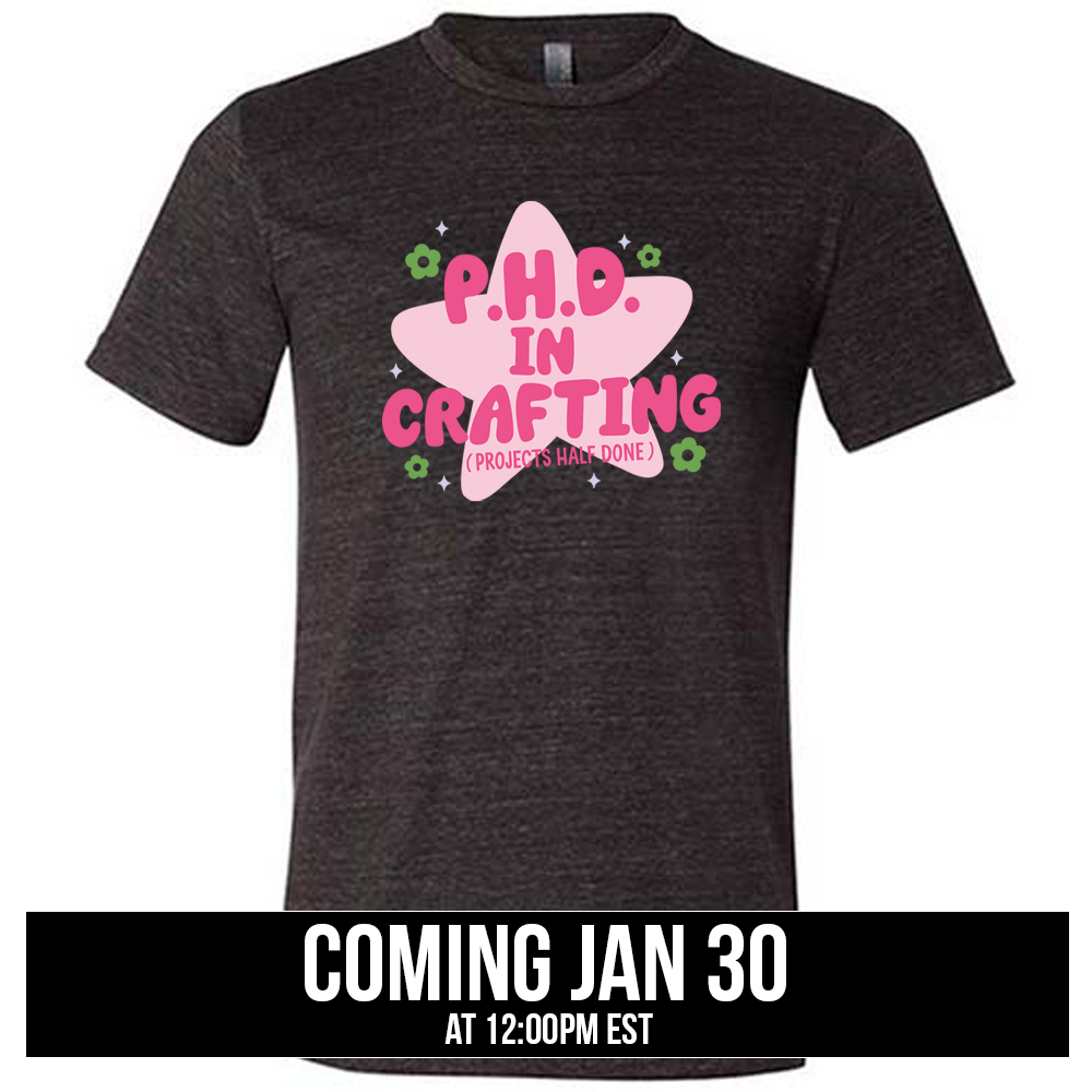 black unisex tee with cartoon graphic of a large star behind the words "P.H.D. in Crafting" in large font with the words (Projects Half Done) in small font underneath, with very small flowers and stars around, coming soon