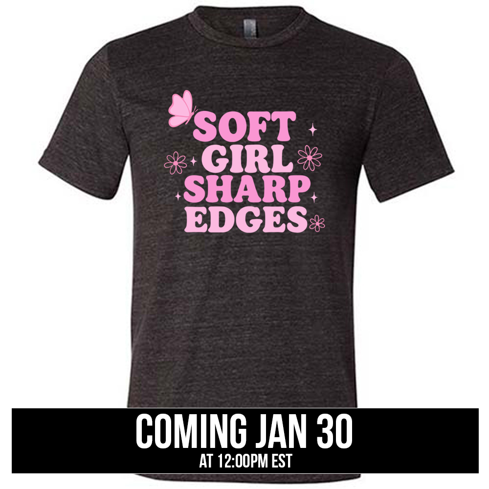 black unisex tee with the words "soft girl sharp edges" in all caps, with graphics of a few little flowers and stars and one butterfly , coming soon