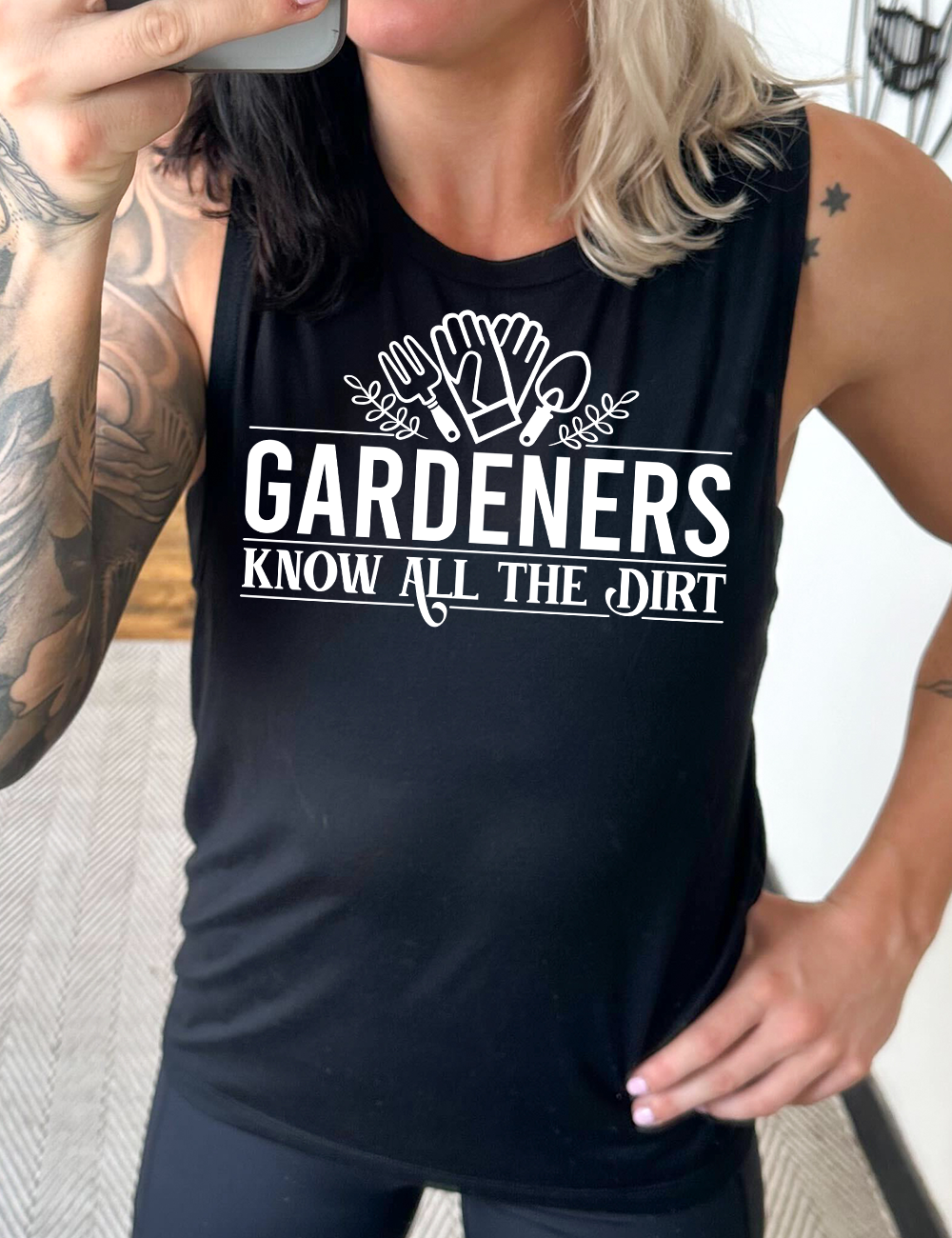 black muscle tank with the words "gardeners know all the dirt" on it, with the graphic of two gloves and two tools above the words