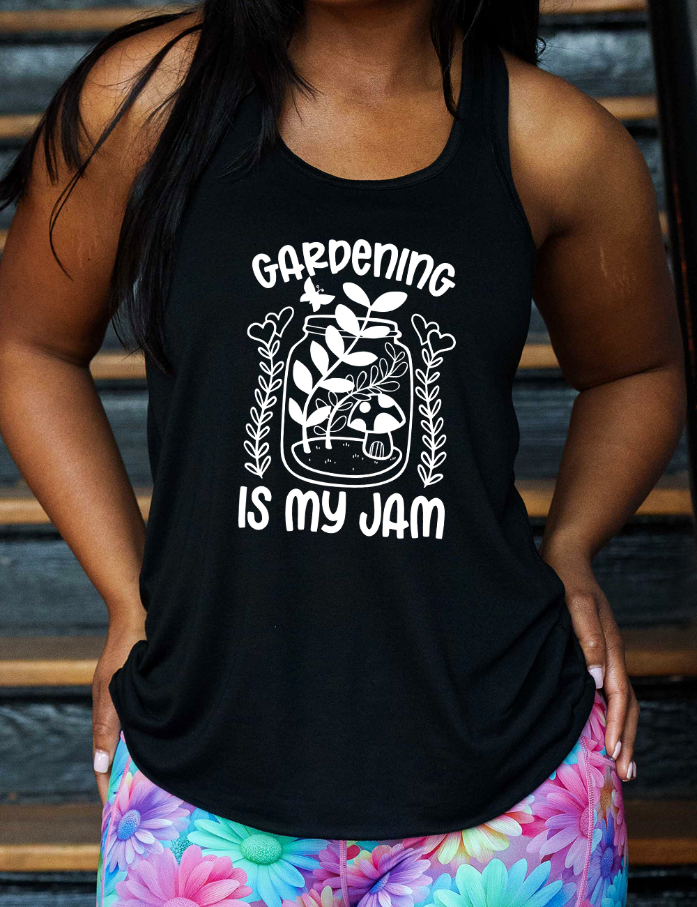black flowy racerback tank with the words "gardening is my jam" on it, and the graphic of a jar with plants and a mushroom in it, a little butterfly flying above it, and a plant (with hearts for flowers on them) on both sides of it
