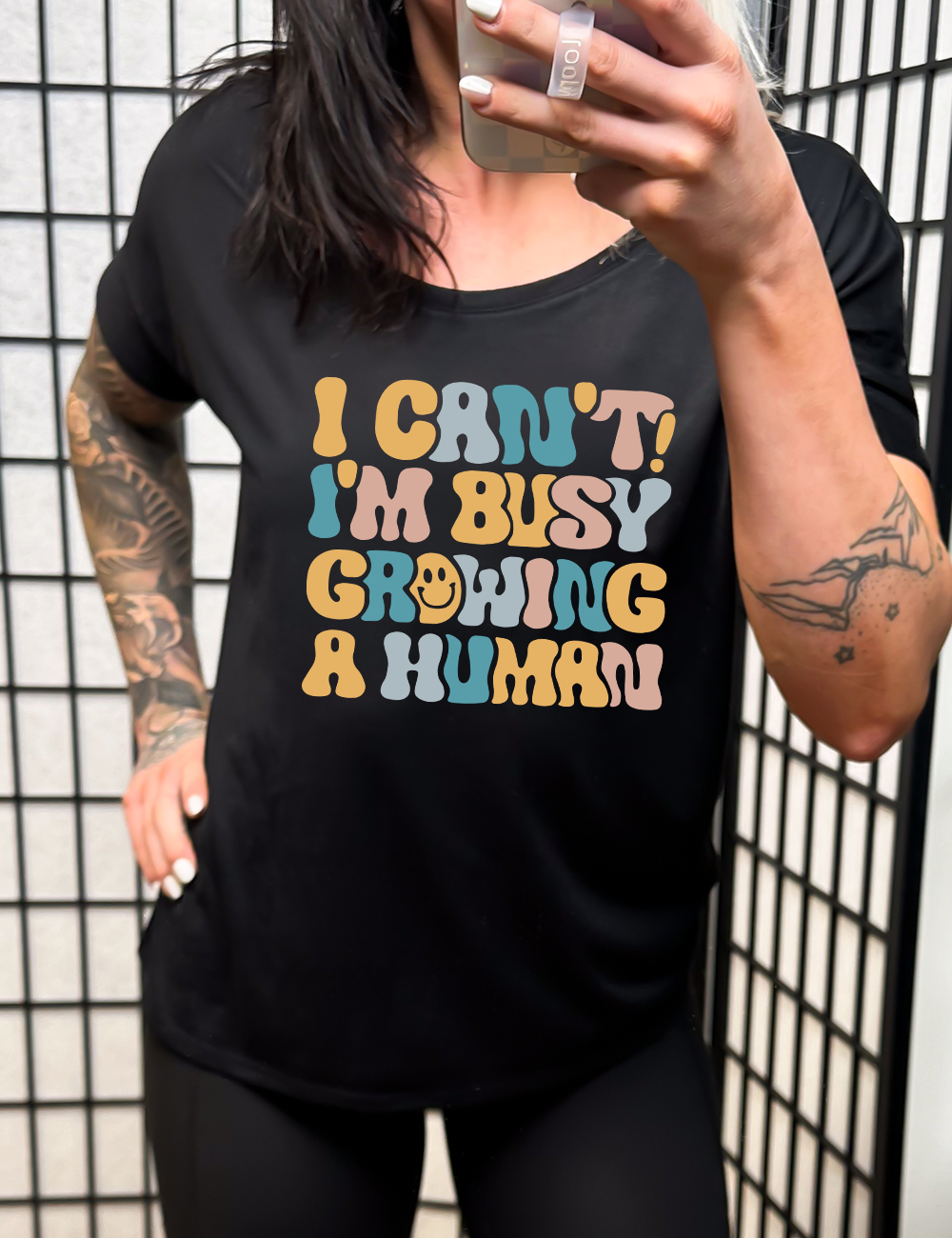 black slouchy tee with the words "I can't! I'm busy growing a human" on it in colorful font, with a smiley face in the "o" of the word "growing"