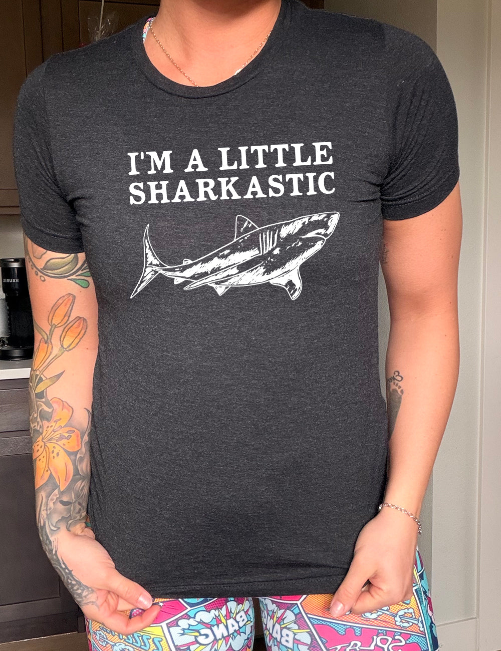 black unisex tee with the words "I'm a little sharkastic" on it, over the graphic of a shark