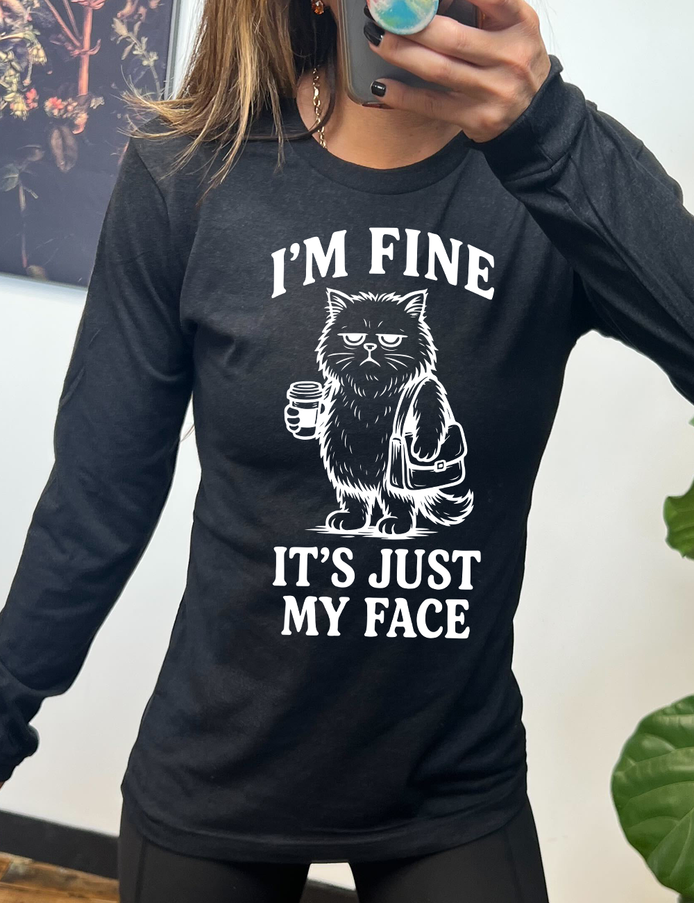 heather black long sleeve shirt with the words "I'm fine it's just my face" on it, around the graphic of a cat with a resting-grumpy face, holding a coffee and wearing a purse