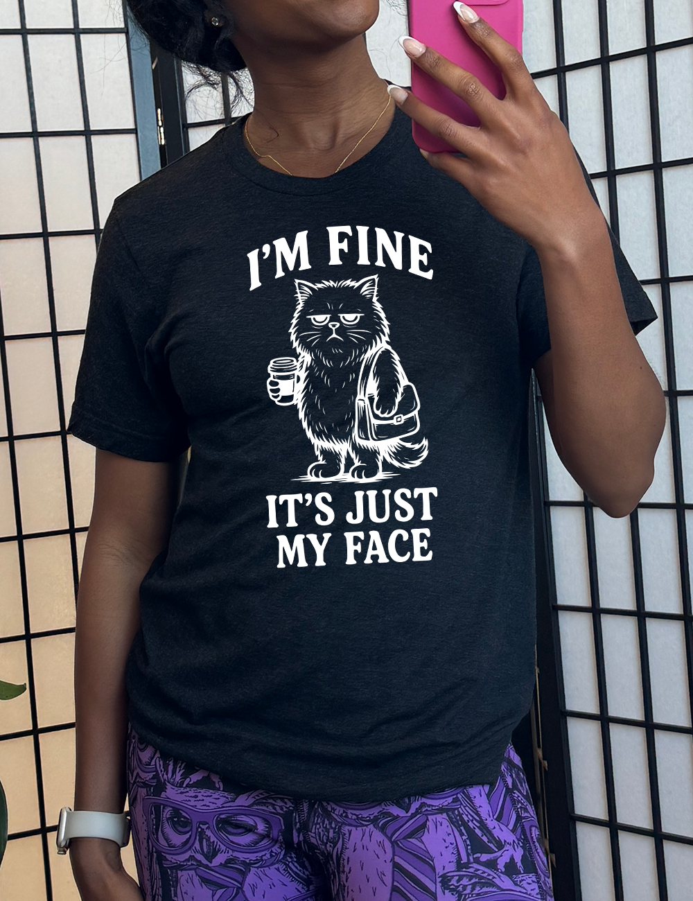 black unisex tee with the words "I'm fine it's just my face" on it, around the graphic of a cat with a resting-grumpy face, holding a coffee and wearing a purse