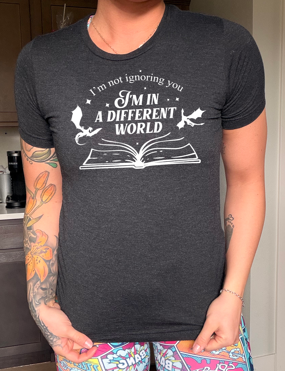 black unisex tee with the words "I'm not ignoring you I'm in a different world" on it, with the graphic of a large book, lying open, and two little dragons flying next to the words, a few stars scattered around