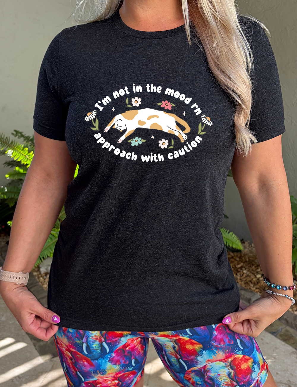 black unisex tee with the words "I'm not in the mood rn approach with caution" on it in an oval, with the graphic of a cat splayed on its back within the words, and a few flowers around