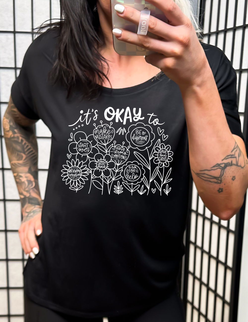black slouchy tee with the words "it's okay to" on it, and underneath there are many flowers that each have the completion of that phrase; the different endings are "make mistakes", "be different", "have bad days", "ask for help", "have strong emotions", "change your mind", "Start over", and "not know everything".