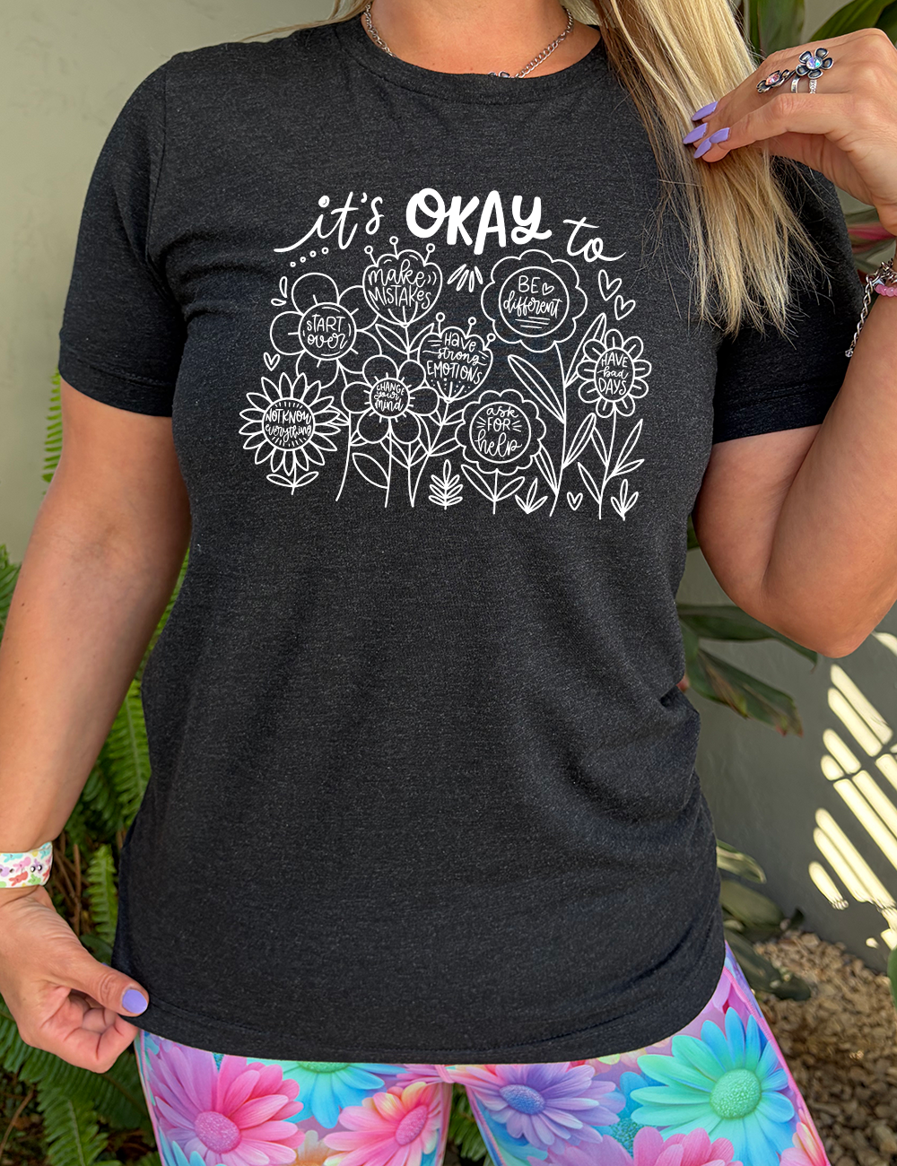 black unisex tee with the words "it's okay to" on it, and underneath there are many flowers that each have the completion of that phrase; the different endings are "make mistakes", "be different", "have bad days", "ask for help", "have strong emotions", "change your mind", "Start over", and "not know everything".