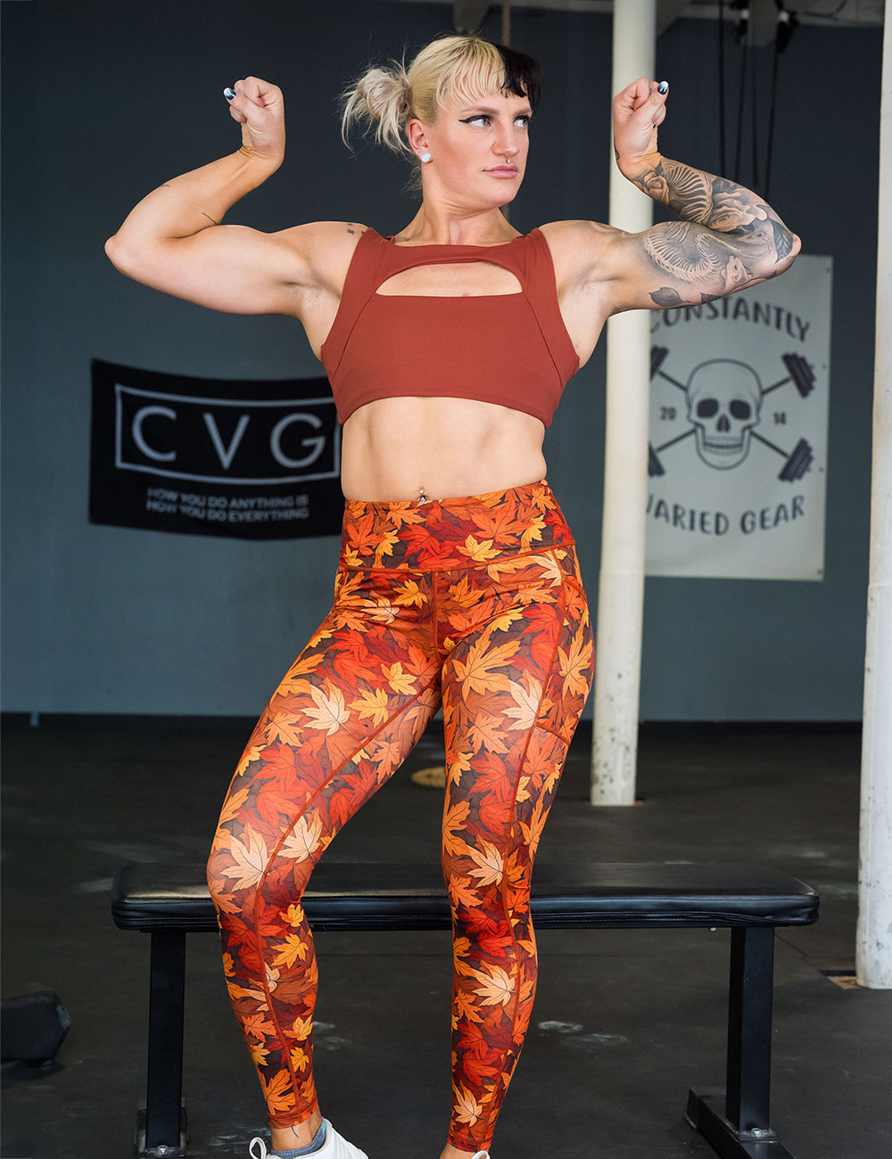 model wearing the orange leaves full length leggings and auburn colored bra flexing in the gym