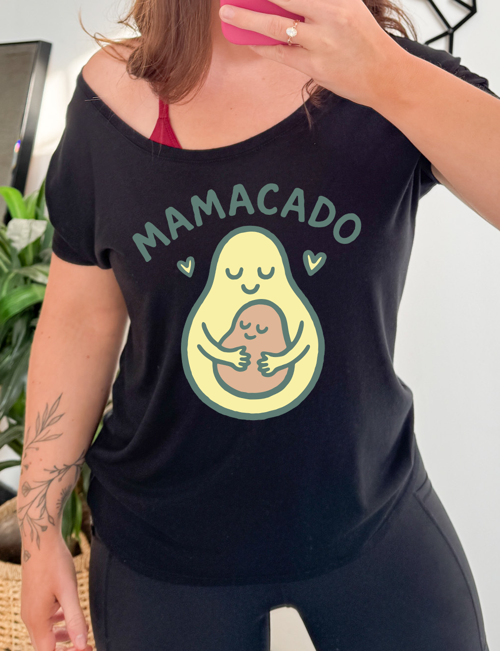 black slouchy tee with the words "mamacado" on it, with the graphic of an avocado, peacefully smiling, with arms, holding her smiling baby (the color and placement of the avocado pit - but in the shape of an avocado), with a little heart on either side