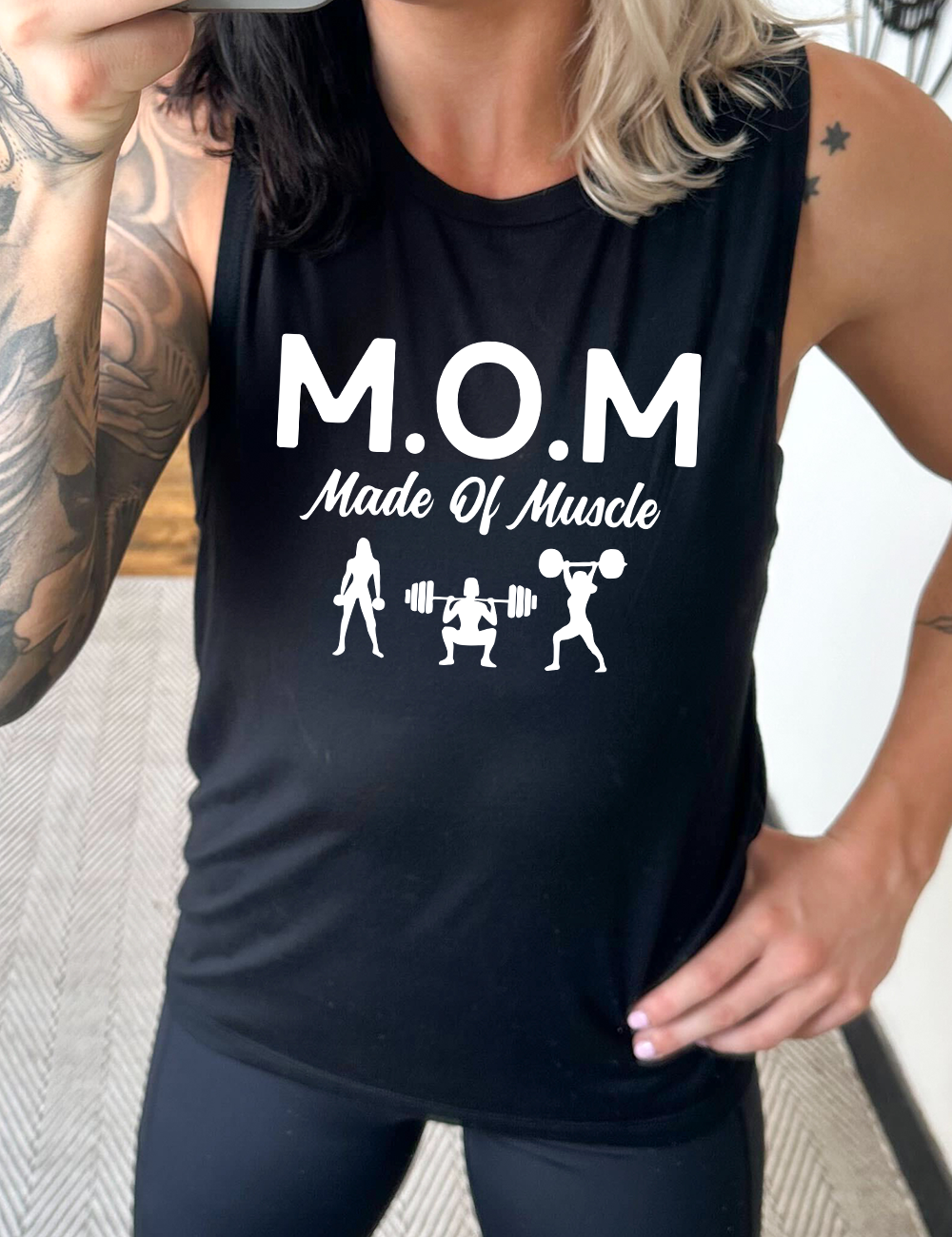 black muscle tank with the word "mom" in large letters as an acronym for the smaller words below it saying "made of muscle", and the graphic of three female silhouettes holding weights