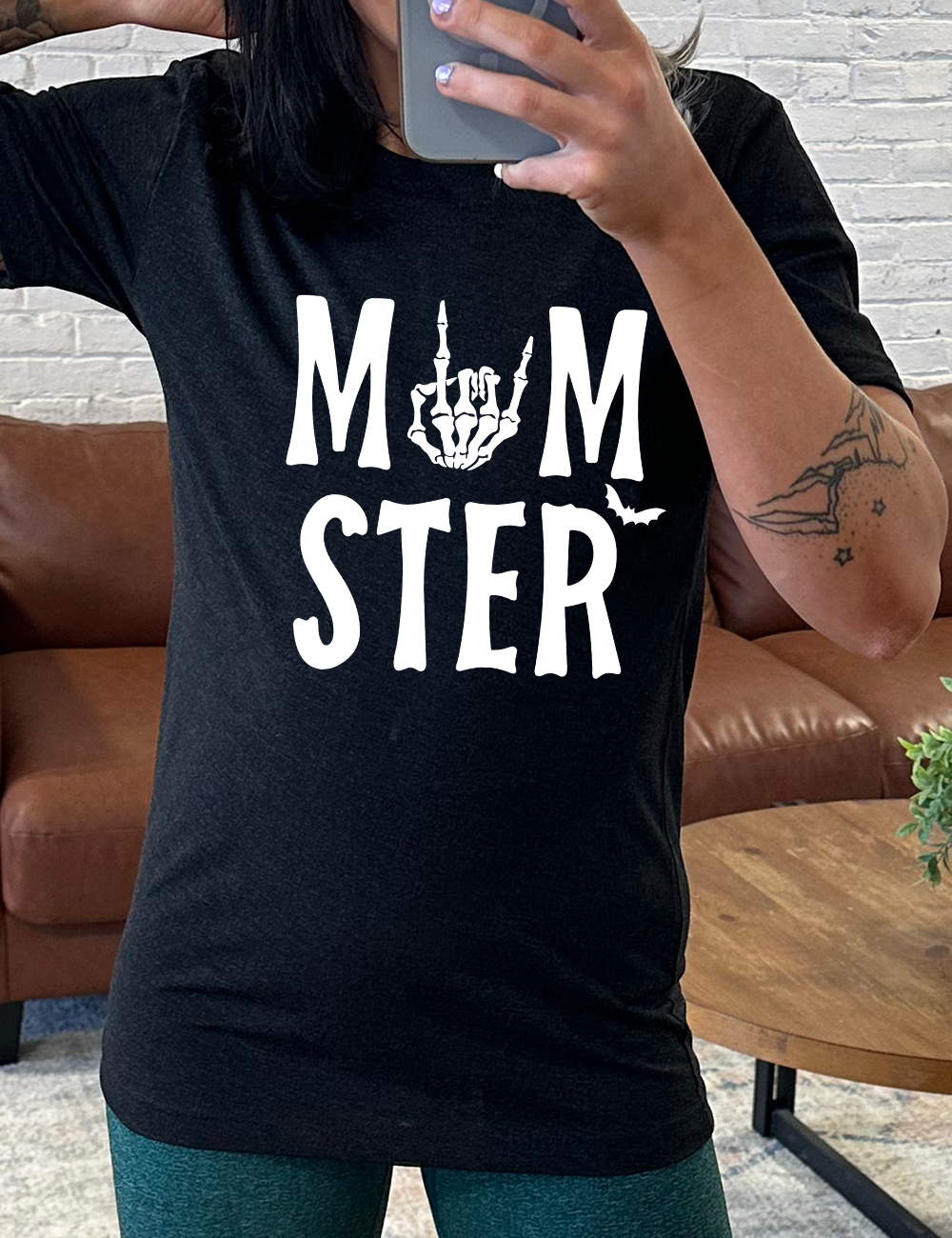 black unisex tee with the word "momster" on it, with the "o" of the word being the graphic of a skeletal rocker hand, and one little bat after the word
