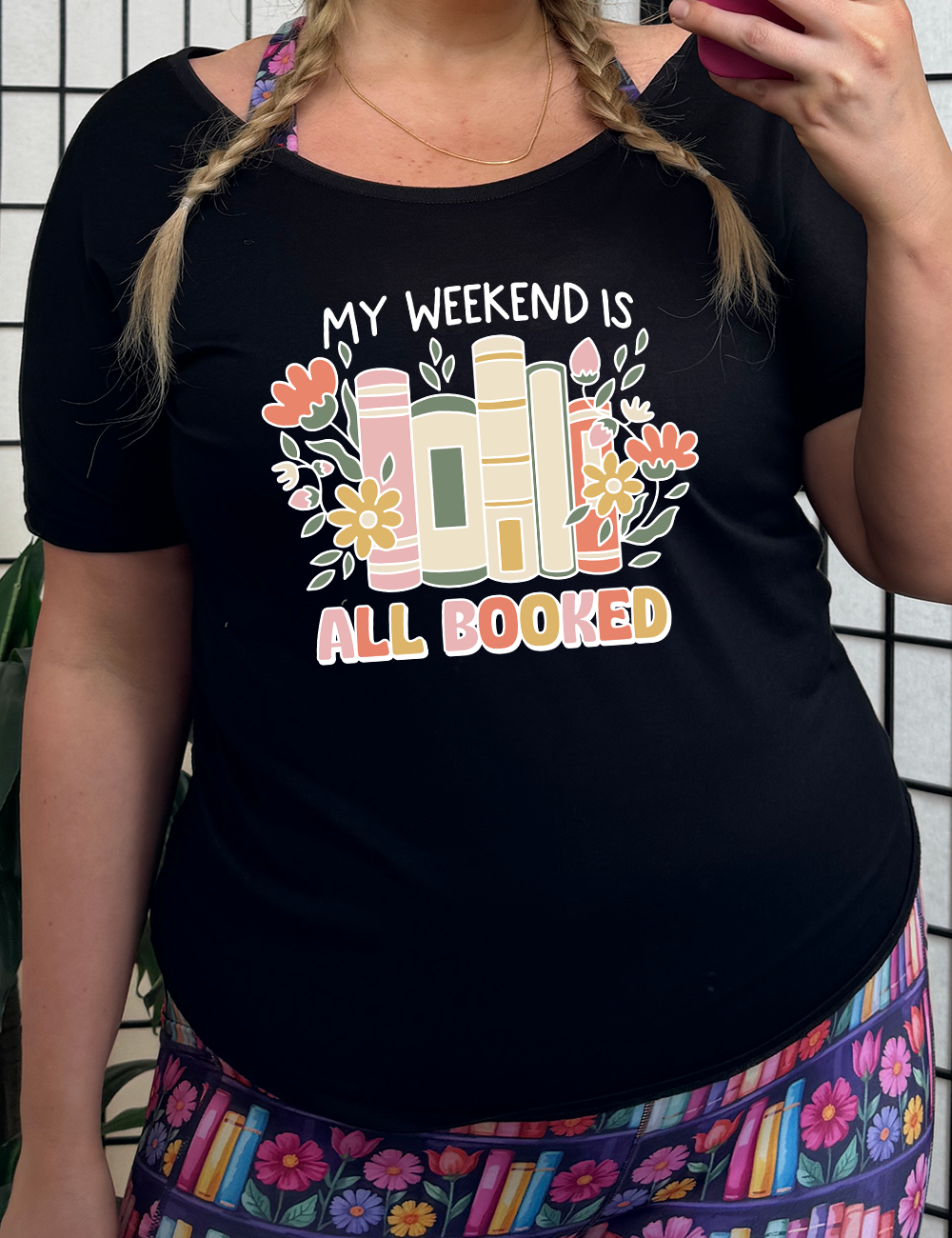A person wearing a black slouchy tee with the text 'MY WEEKEND IS ALL BOOKED' on the front, decorated with images of books and flowers.