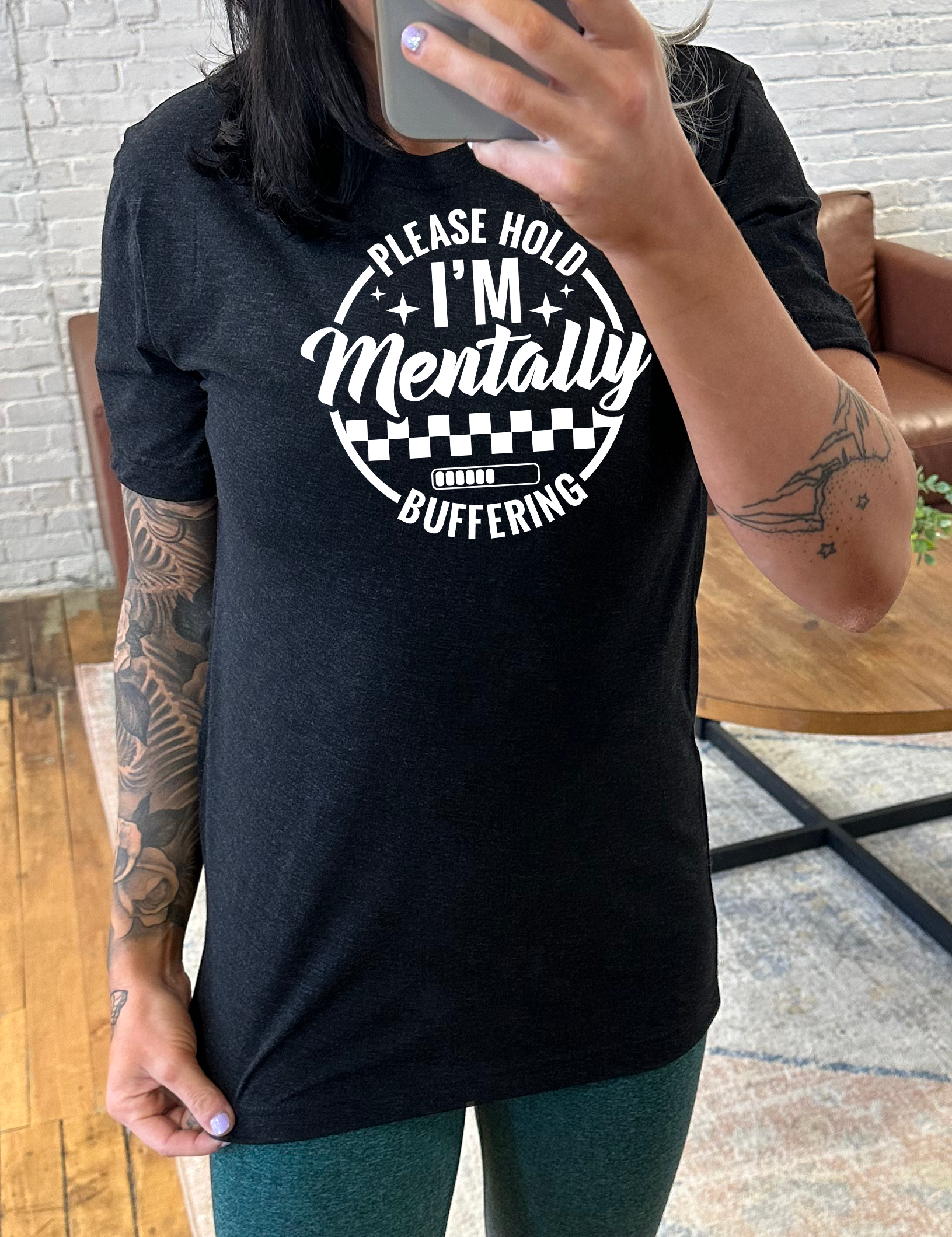black unisex tee with the words "please hold I'm mentally buffering" written on it in a circle graphic, with some of the words part of the arches, and within the circle there are two layers of checkerboard as well as a buffering bar, which is about 6 out of 10 full