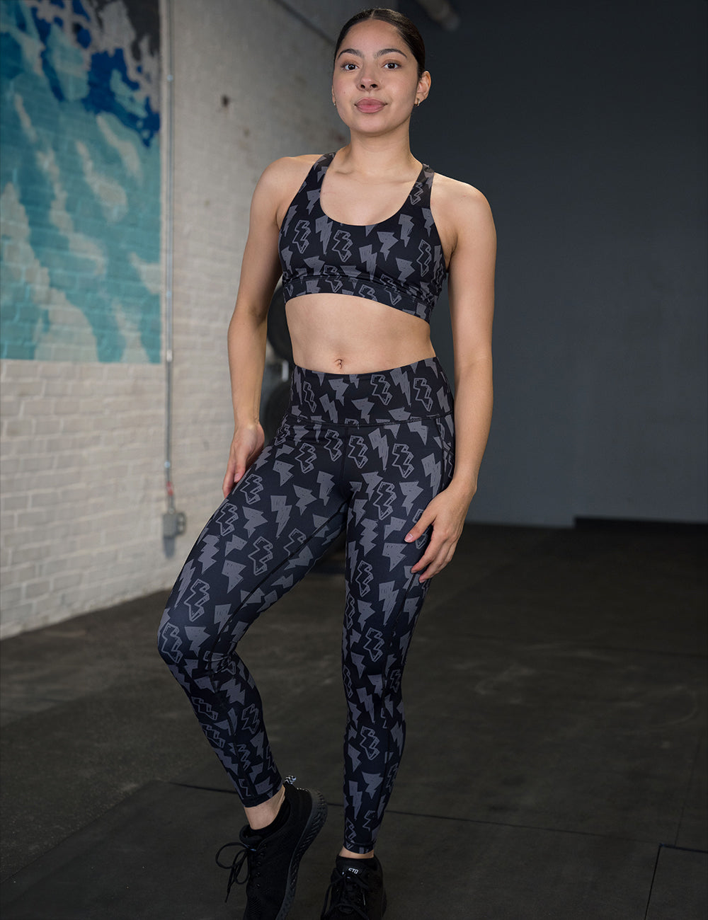 model wearing the full length black leggings with a grey thunderbolts pattern posing in the gym