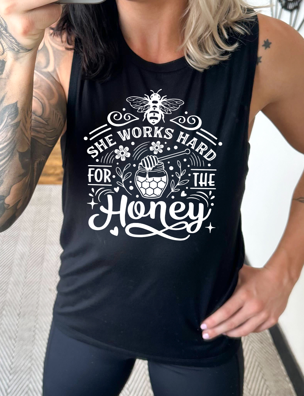 black muscle tank with the words "she works for the honey" surrounded by accenting curved lines, with flowers, dots, stars, and hears scattered around, and a honeypot and spoon in the center, and a bee on the top