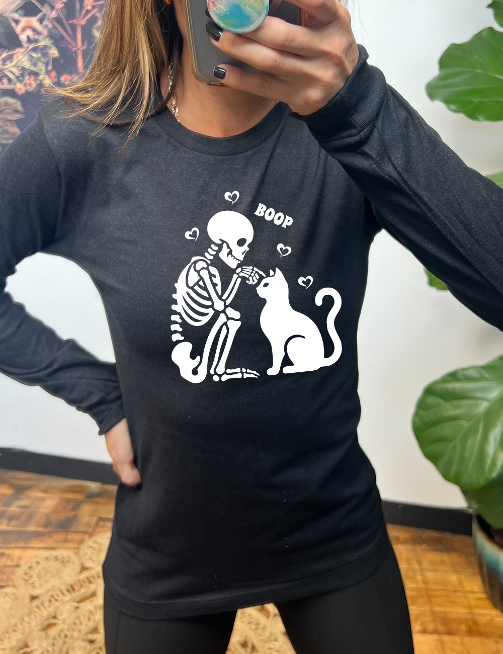 heather black long sleeve shirt with the graphic of a sitting skeleton touching the head of a sitting cat, with the word "boop" above this action, and four little hearts around them