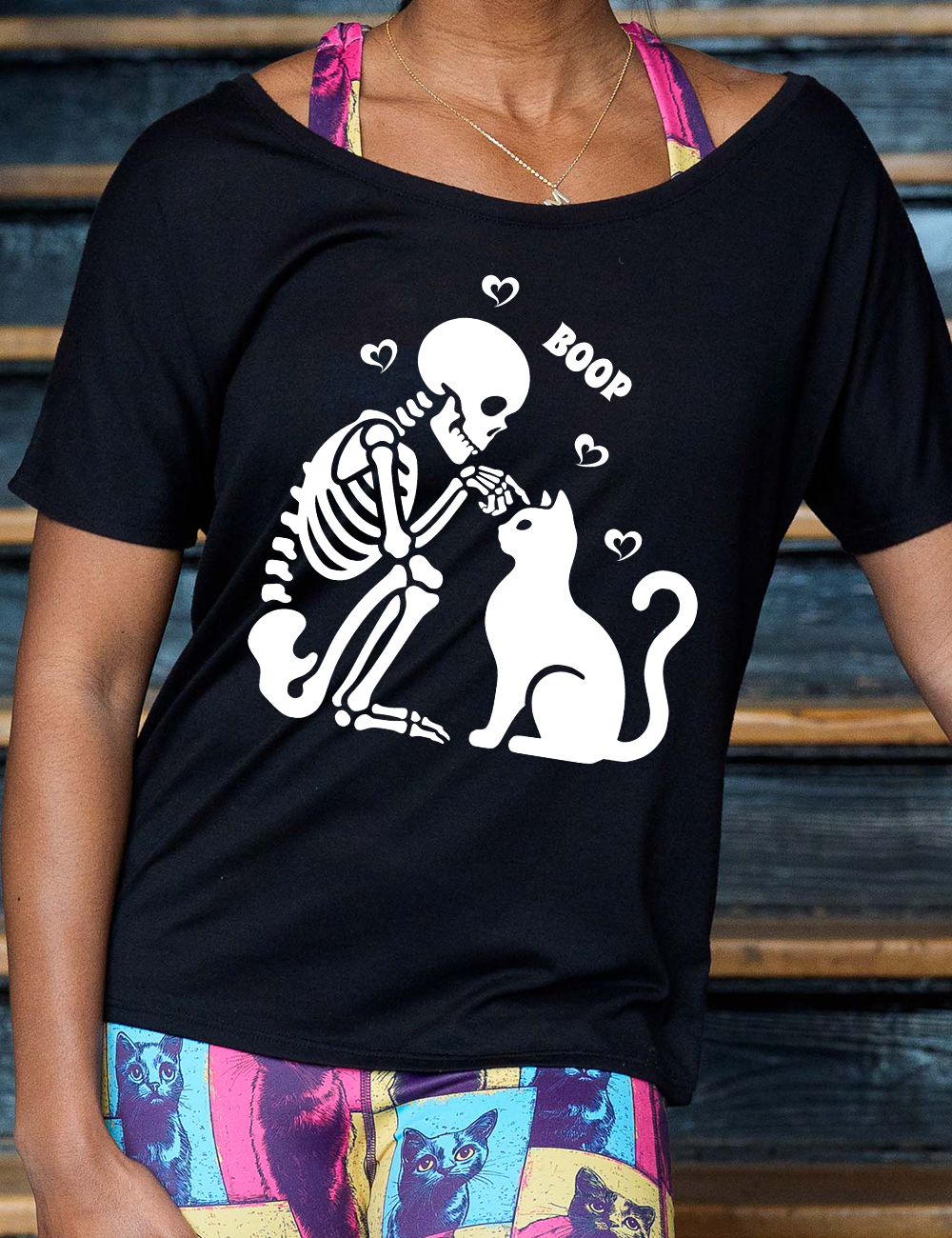 black slouchy tee with the graphic of a sitting skeleton touching the head of a sitting cat, with the word "boop" above this action, and four little hearts around them