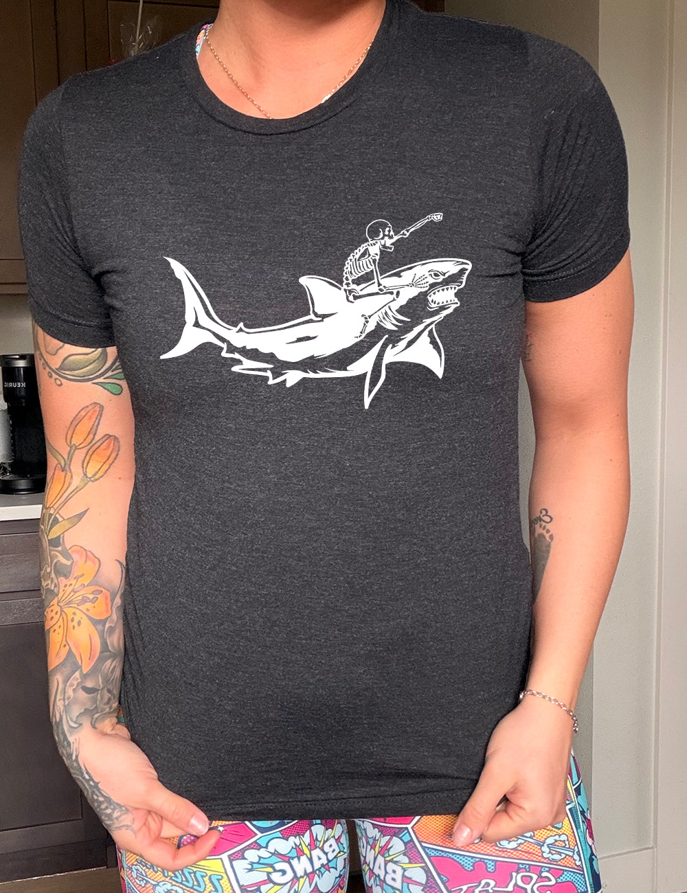 black unisex tee with the graphic of a skeleton, with its fist pointing onwards and its mouth open, riding a shark, who has a fierce expression