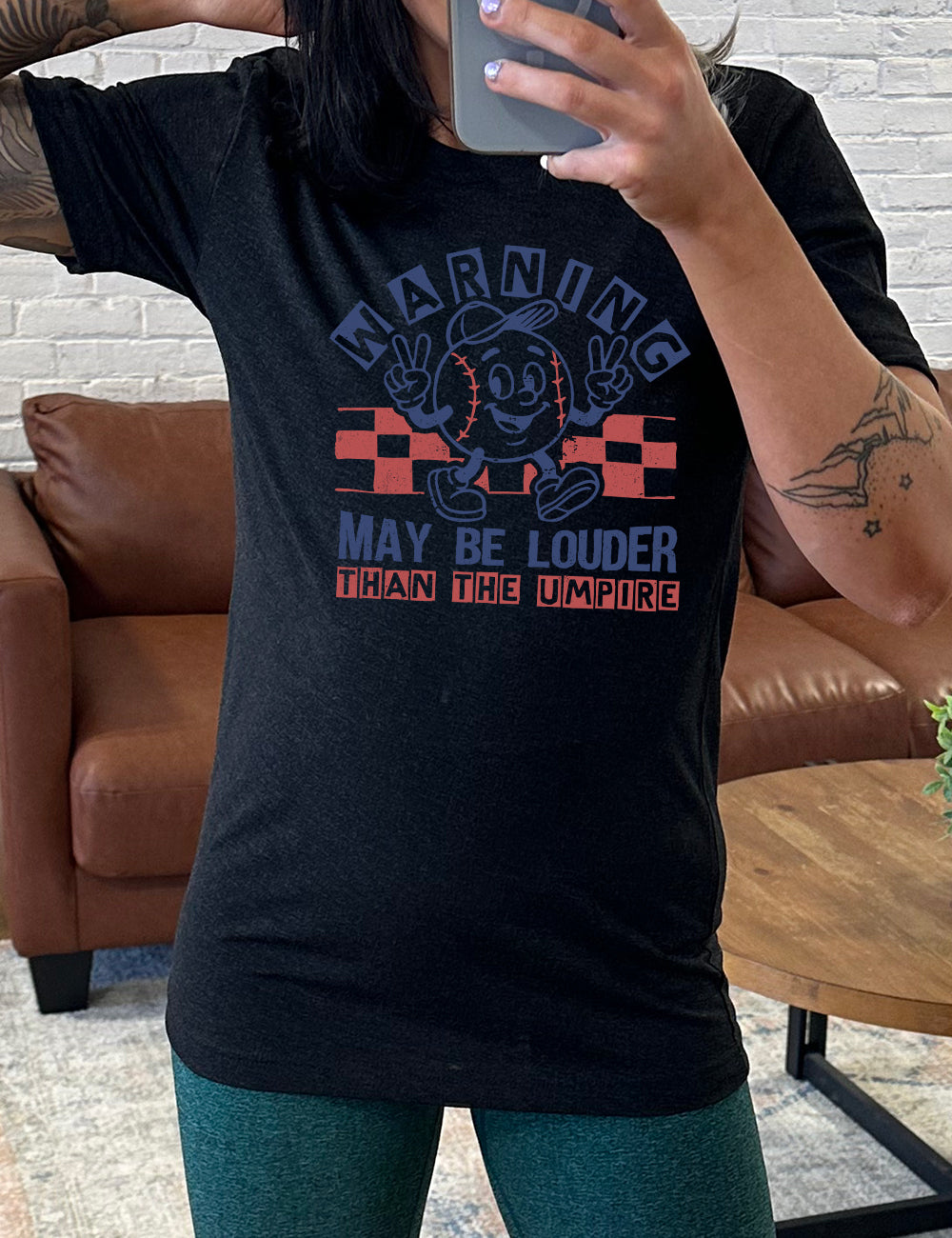black unisex tee with the words "warning may be louder than the umpire" on it, with the graphic of baseball with a face, wearing a hat, holding up two peace signs, and walking along, with a checkered border behind it