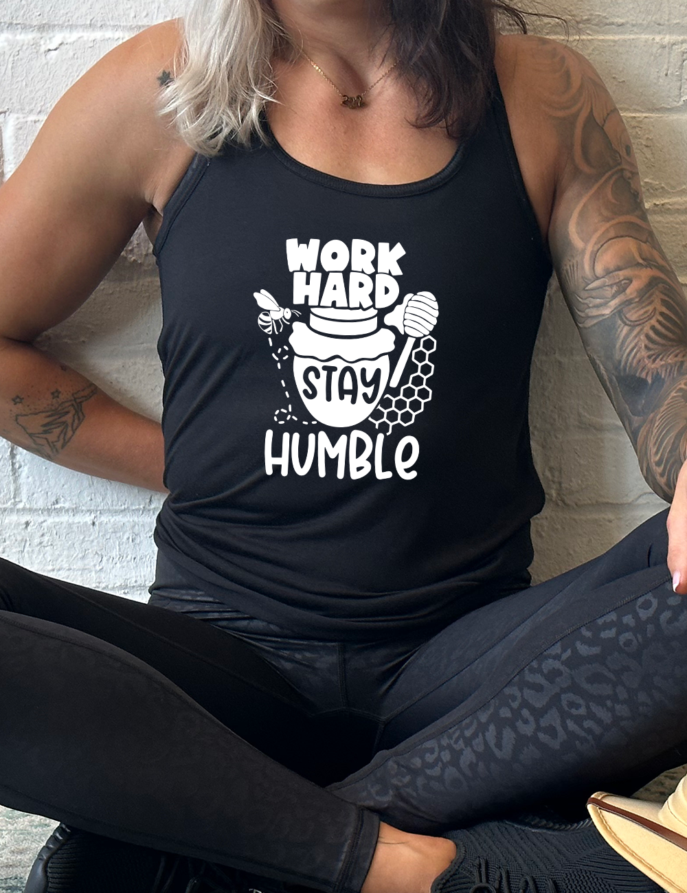 black flowy racerback tank with the words "work hard stay humble" on it, with the word "stay" on the graphic of a honeypot, and a bee flying around the pot's left, and some honeycomb and a honey spoon on its right
