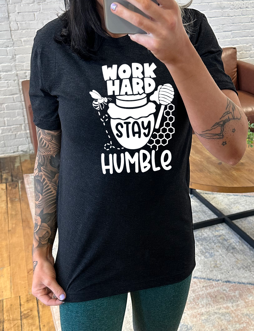 black unisex tee with the words "work hard stay humble" on it, with the word "stay" on the graphic of a honeypot, and a bee flying around the pot's left, and some honeycomb and a honey spoon on its right