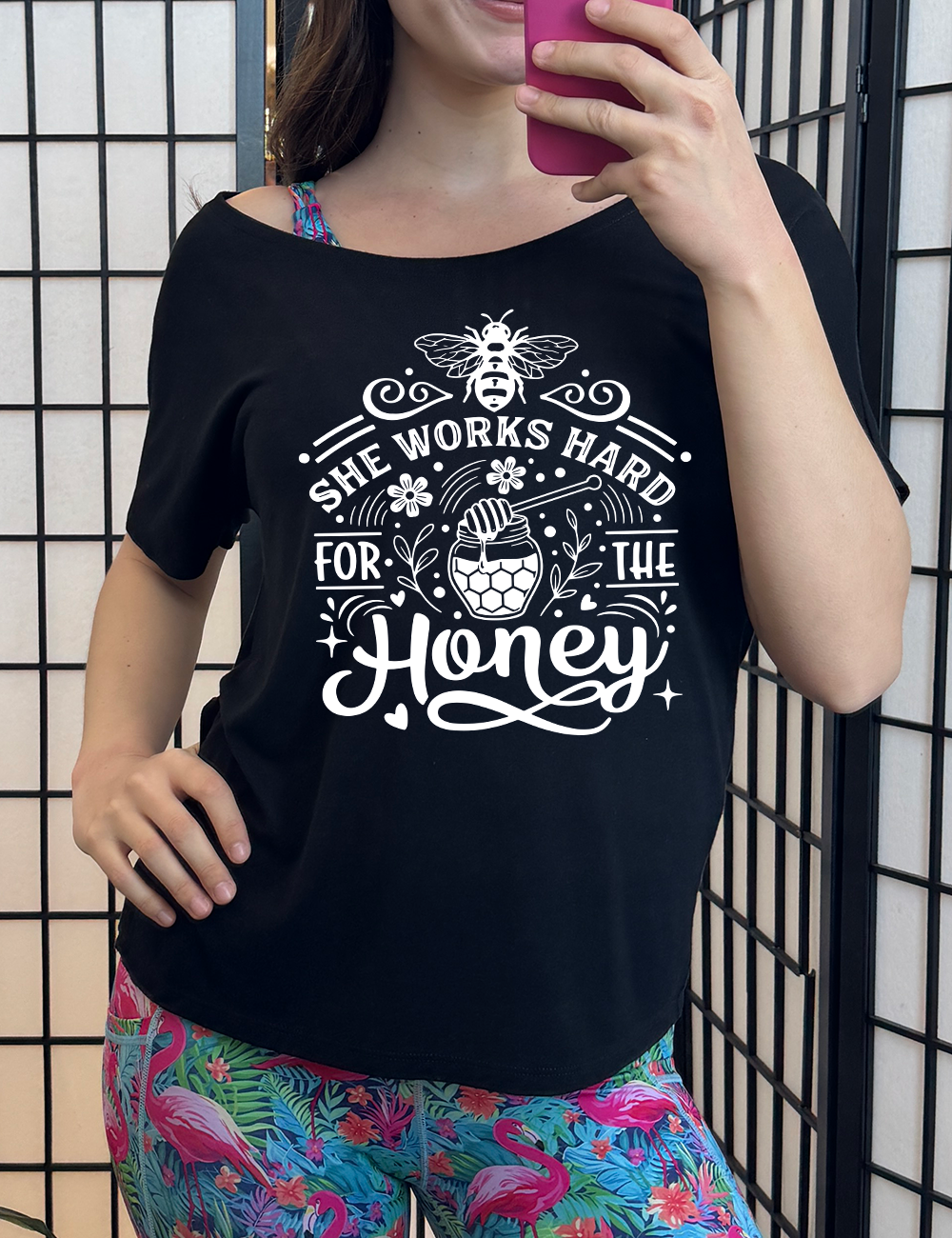 black slouchy tee with the words "she works for the honey" surrounded by accenting curved lines, with flowers, dots, stars, and hears scattered around, and a honeypot and spoon in the center, and a bee on the top