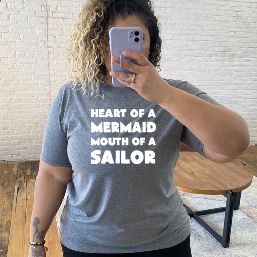 Model wearing a grey unisex tee with the saying "heart of a mermaid mouth of a sailor" in white