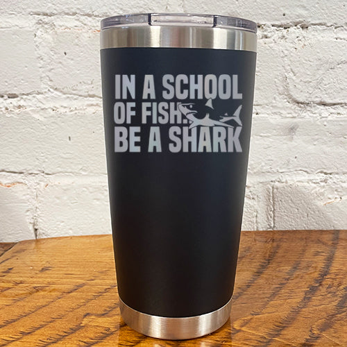 20oz black tumbler with silver saying "in a school of fish be a shark" with a shark cartoon over the word "fish"
