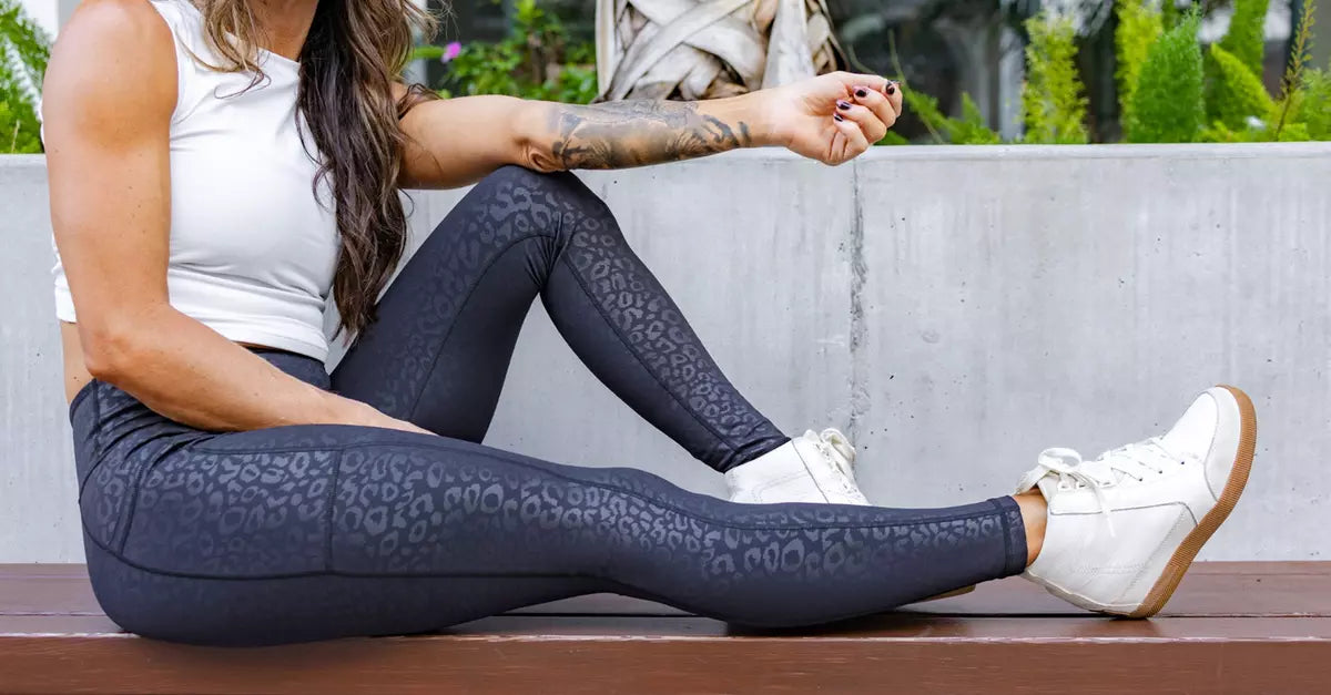 Woman sitting on a bench wearing patterned black leggings and white sneakers outdoors.