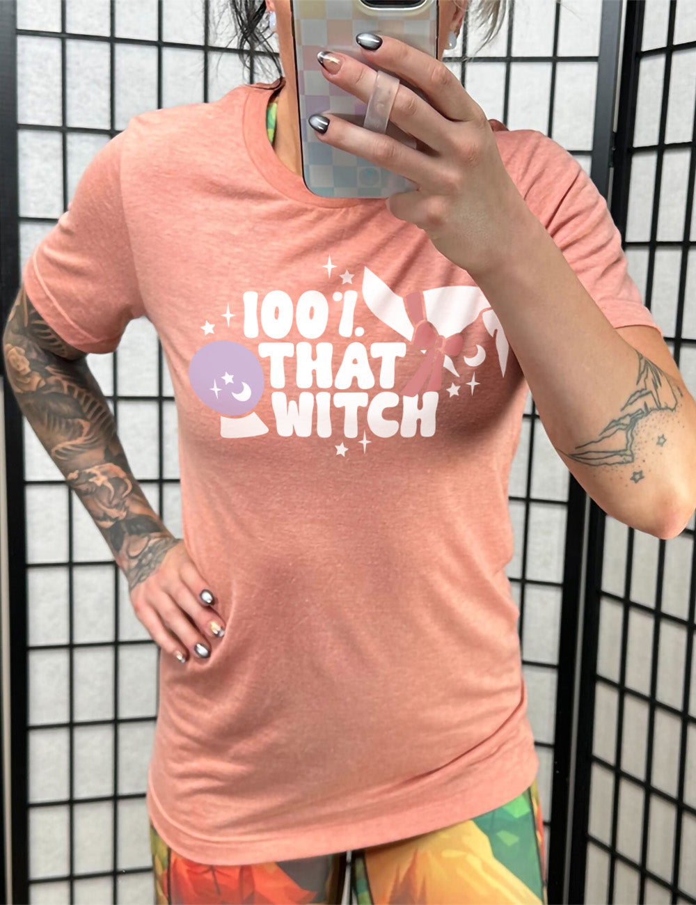 sunset unisex tee with the words "100% that witch" next to the graphic of a witch hat with a bow, a crystal ball, and some stars