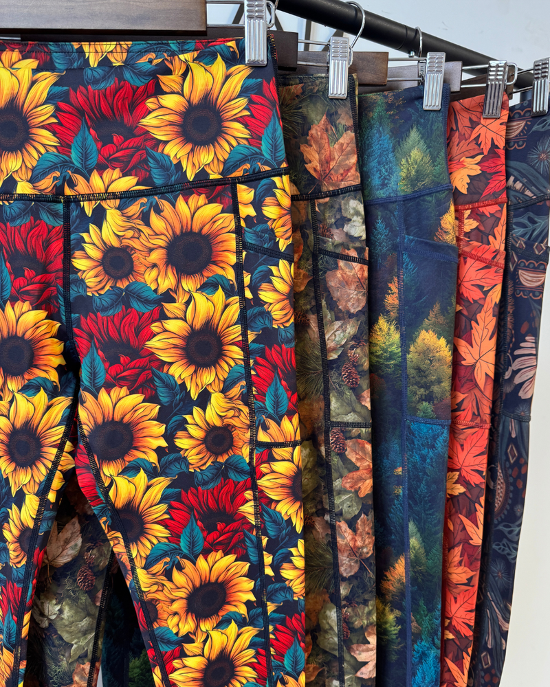 A close-up of several pairs of leggings hanging on a black rack with metal clips. The front pair has a bold sunflower and red floral print on a dark background, while the adjacent pairs show different autumn-themed prints including leaves and foliage in teal, orange, and brown tones.
