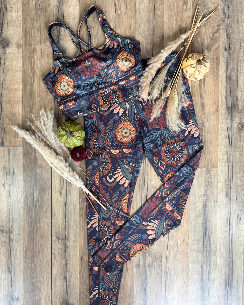 A matching two-piece activewear set featuring a bohemian-style geometric and floral mandala print in navy, rust orange, and beige, styled flat-lay on a light wood floor with dried pampas grass and small decorative pumpkins.
