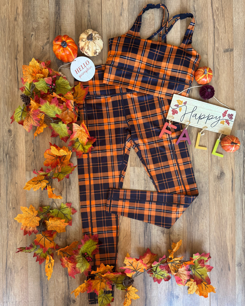 A plaid outfit set styled in an autumn flat lay on a wooden floor with fall leaves, pumpkins, and a 'Happy Fall' sign.
