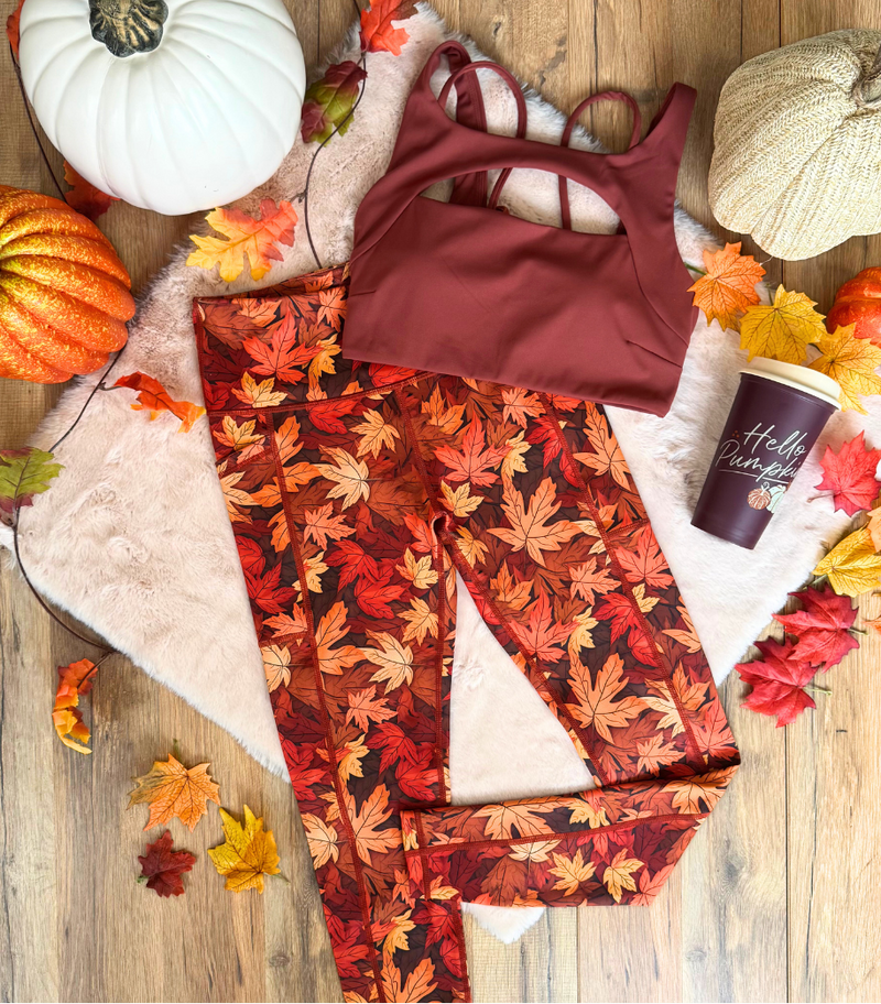 A styled flat-lay of a two-piece activewear set featuring a rust-brown sports bra and matching leggings with a red, orange, and brown autumn leaf pattern, arranged on a wooden floor with pumpkins, faux leaves, a cup, and a blanket.