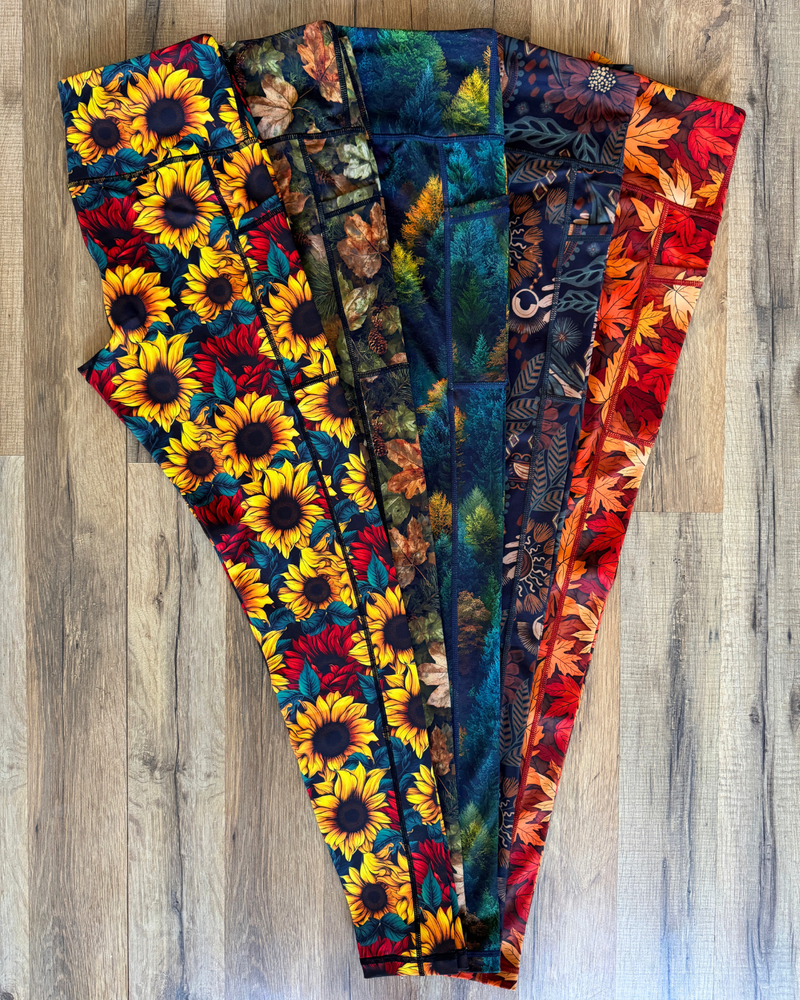 Five pairs of leggings with different autumn and floral prints are fanned out on a light brown wood floor. 
