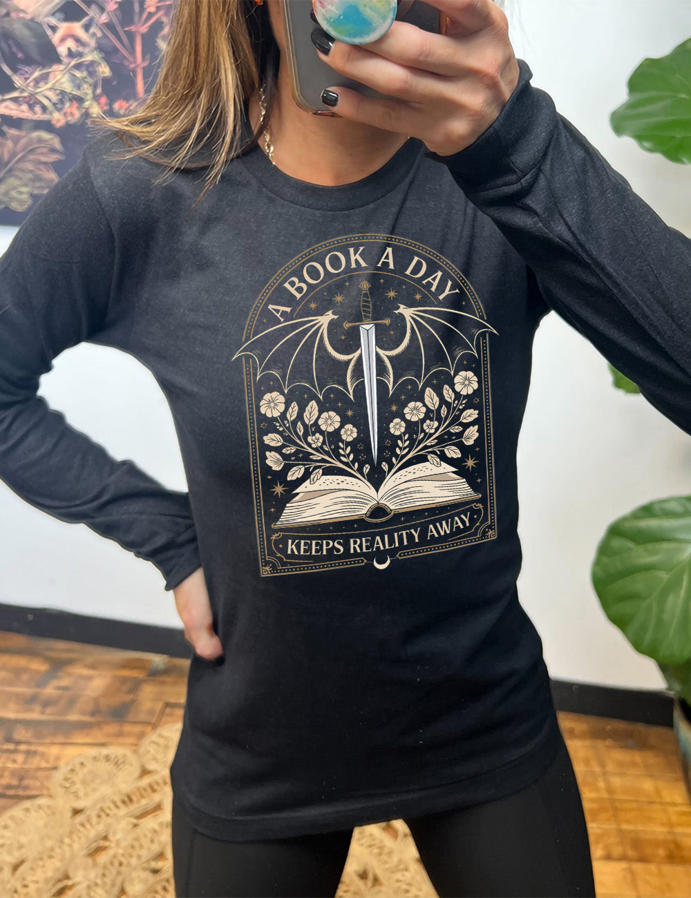 heather black long sleeve shirt with the graphic of an archway, and within it, the graphic of an open book, with flowers growing out of it, the graphic of a sword poised downwards, with dragon wings, and many stars throughout, and the words saying "a book a day keeps reality away"