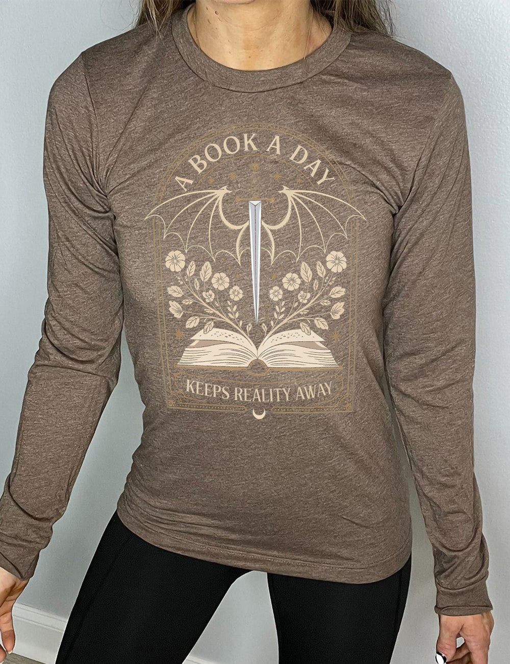 heather brown long sleeve shirt with the graphic of an archway, and within it, the graphic of an open book, with flowers growing out of it, the graphic of a sword poised downwards, with dragon wings, and many stars throughout, and the words saying "a book a day keeps reality away"