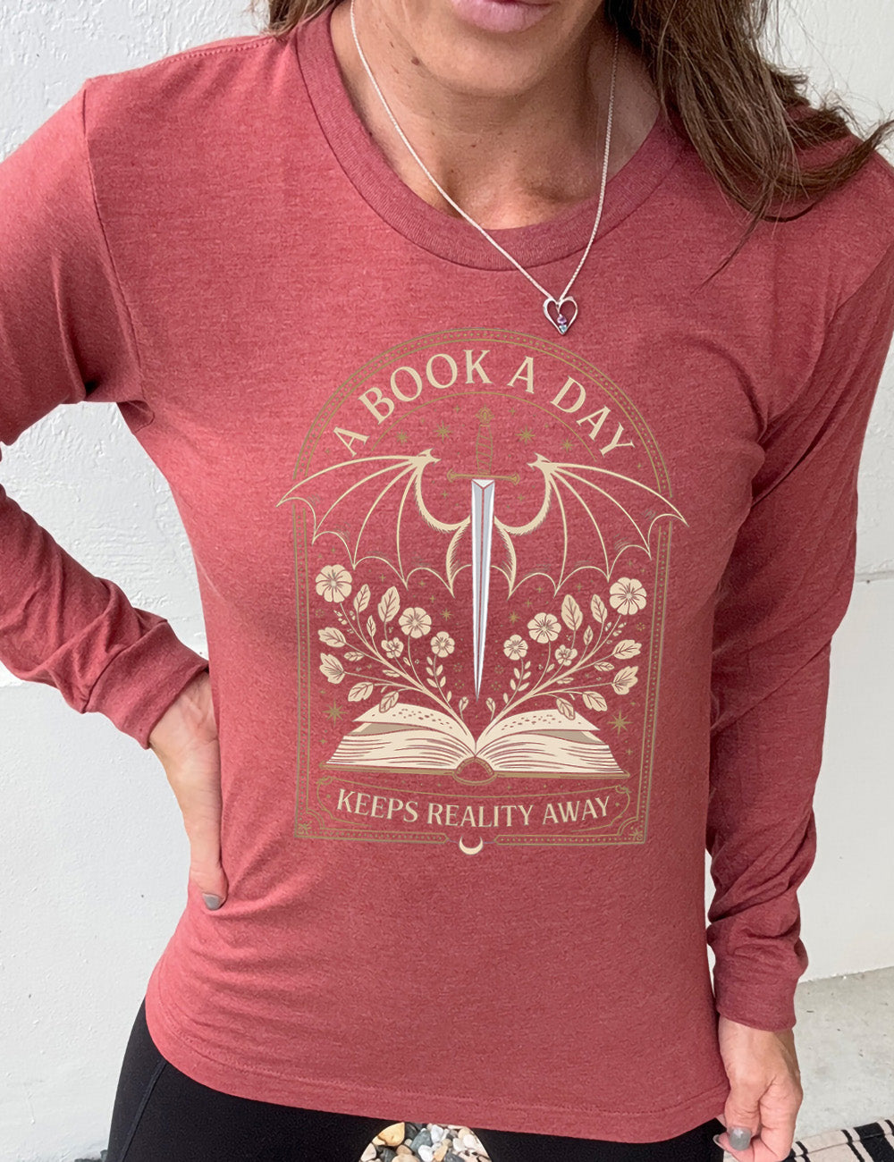 heather clay long sleeve shirt with the graphic of an archway, and within it, the graphic of an open book, with flowers growing out of it, the graphic of a sword poised downwards, with dragon wings, and many stars throughout, and the words saying "a book a day keeps reality away"
