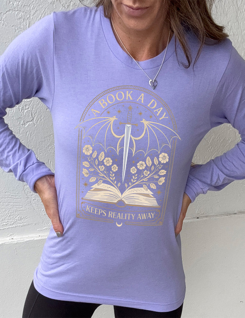 heather dark lavender long sleeve shirt with the graphic of an archway, and within it, the graphic of an open book, with flowers growing out of it, the graphic of a sword poised downwards, with dragon wings, and many stars throughout, and the words saying "a book a day keeps reality away"