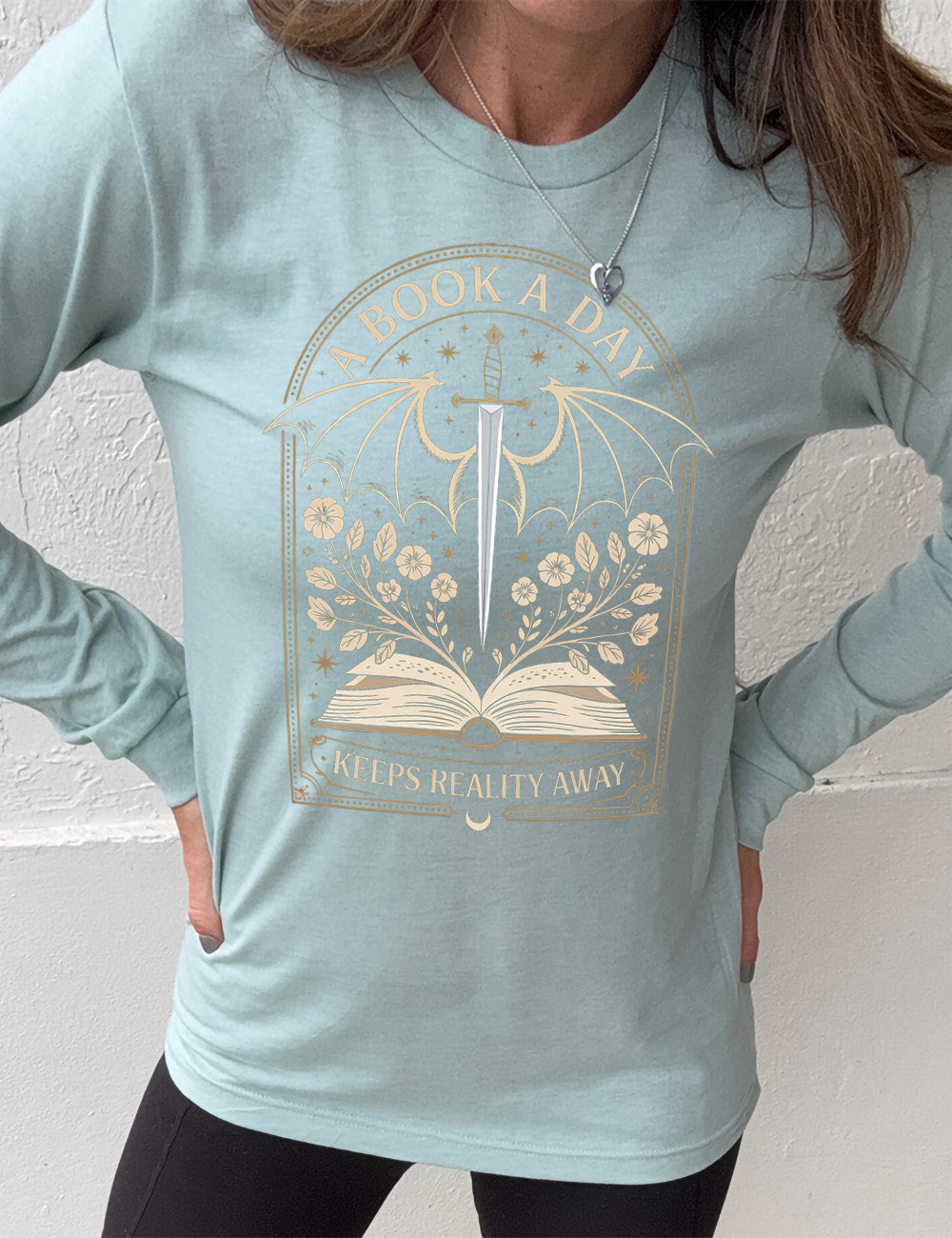 heather dusty blue long sleeve shirt with the graphic of an archway, and within it, the graphic of an open book, with flowers growing out of it, the graphic of a sword poised downwards, with dragon wings, and many stars throughout, and the words saying "a book a day keeps reality away"