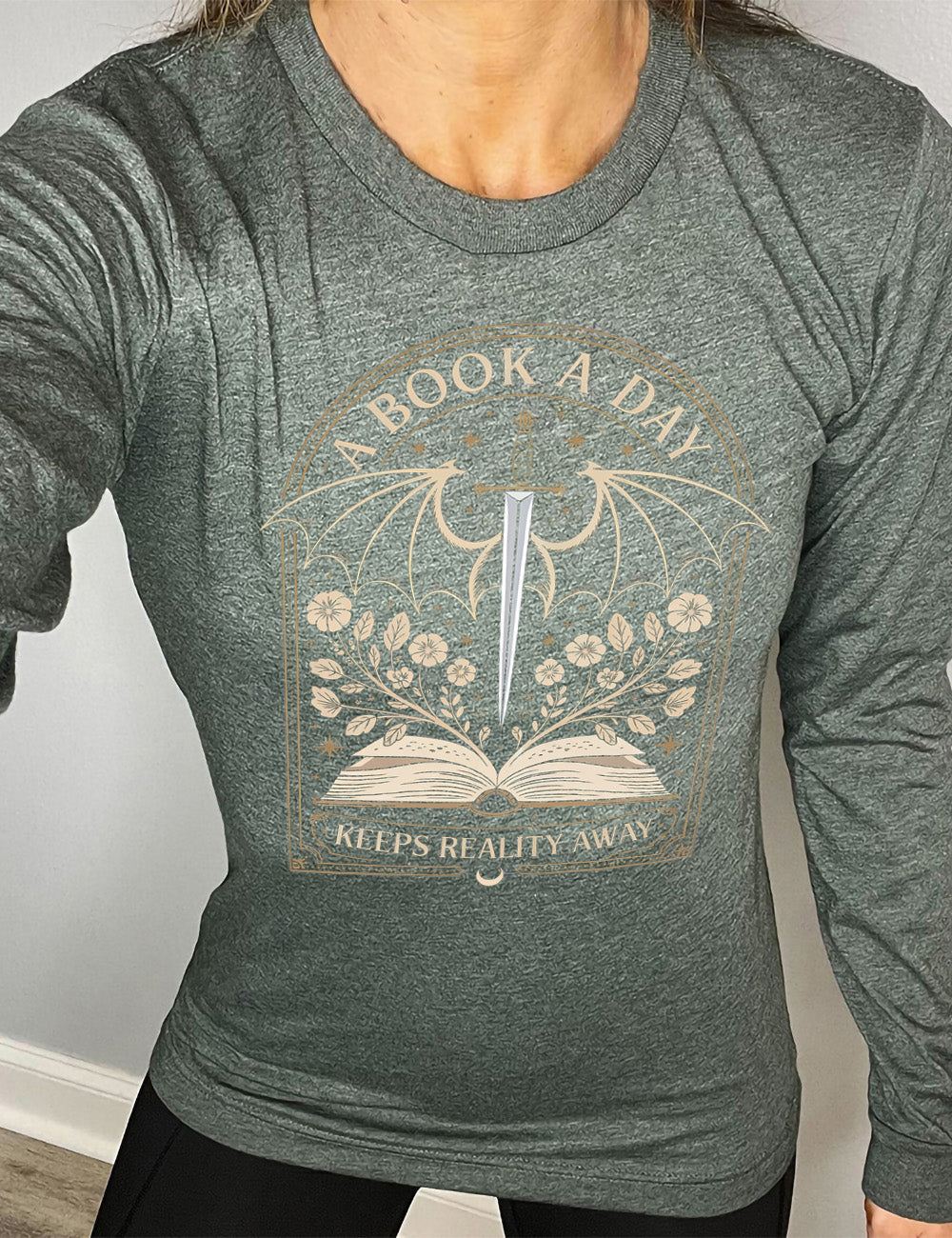 heather forest long sleeve shirt with the graphic of an archway, and within it, the graphic of an open book, with flowers growing out of it, the graphic of a sword poised downwards, with dragon wings, and many stars throughout, and the words saying "a book a day keeps reality away"
