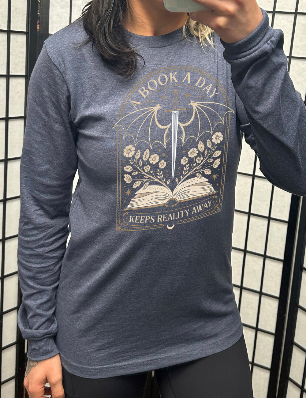 heather navy long sleeve shirt with the graphic of an archway, and within it, the graphic of an open book, with flowers growing out of it, the graphic of a sword poised downwards, with dragon wings, and many stars throughout, and the words saying "a book a day keeps reality away"