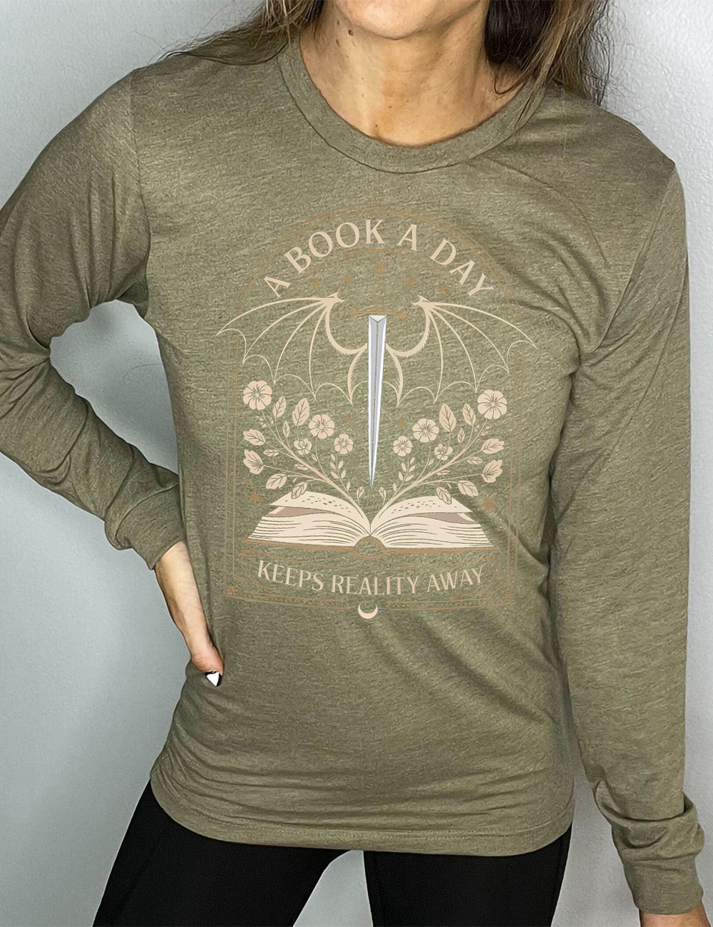 heather olive long sleeve shirt with the graphic of an archway, and within it, the graphic of an open book, with flowers growing out of it, the graphic of a sword poised downwards, with dragon wings, and many stars throughout, and the words saying "a book a day keeps reality away"