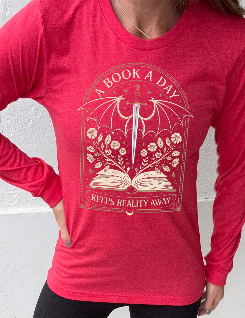 heather red long sleeve shirt with the graphic of an archway, and within it, the graphic of an open book, with flowers growing out of it, the graphic of a sword poised downwards, with dragon wings, and many stars throughout, and the words saying "a book a day keeps reality away"