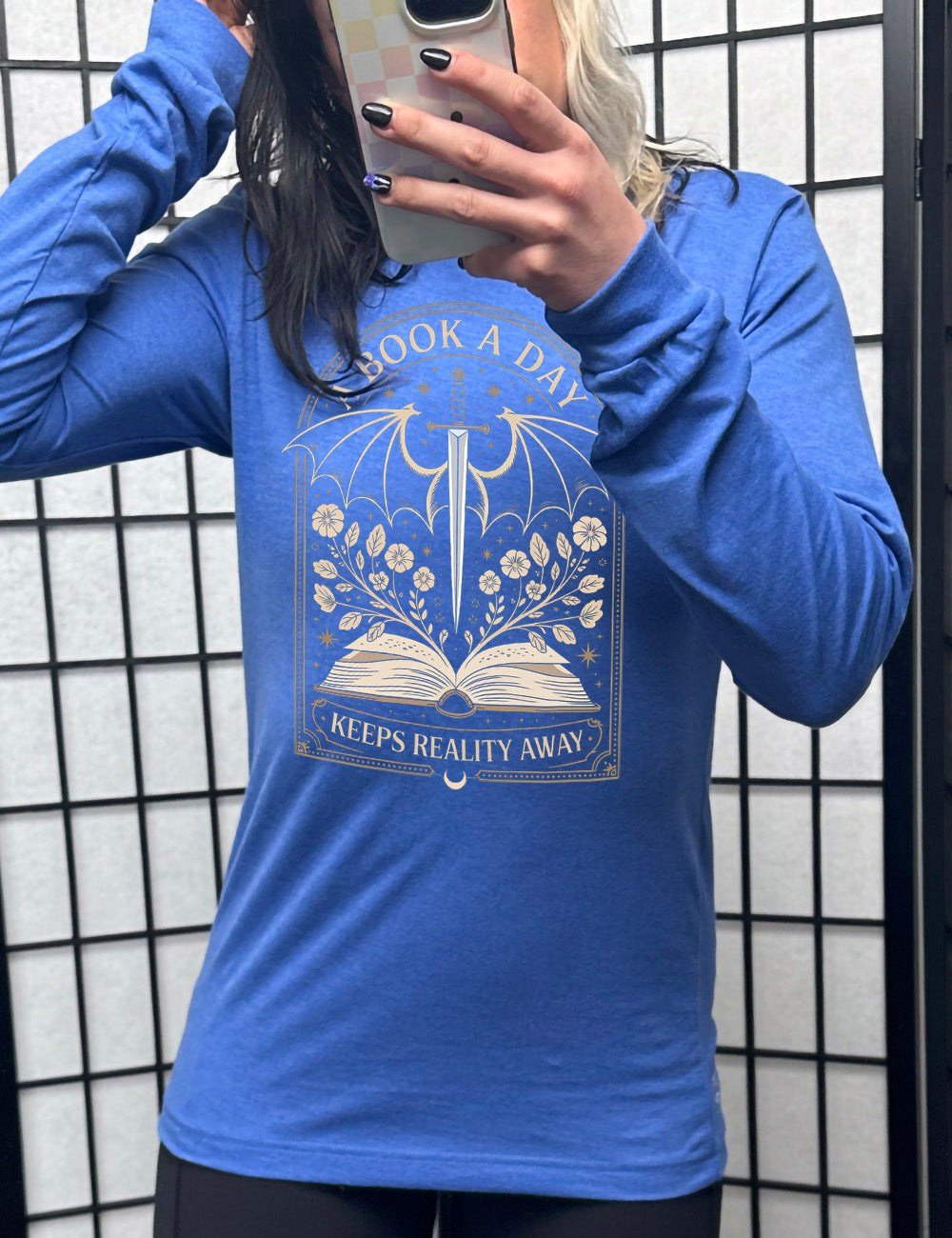heather true royal long sleeve shirt with the graphic of an archway, and within it, the graphic of an open book, with flowers growing out of it, the graphic of a sword poised downwards, with dragon wings, and many stars throughout, and the words saying "a book a day keeps reality away"