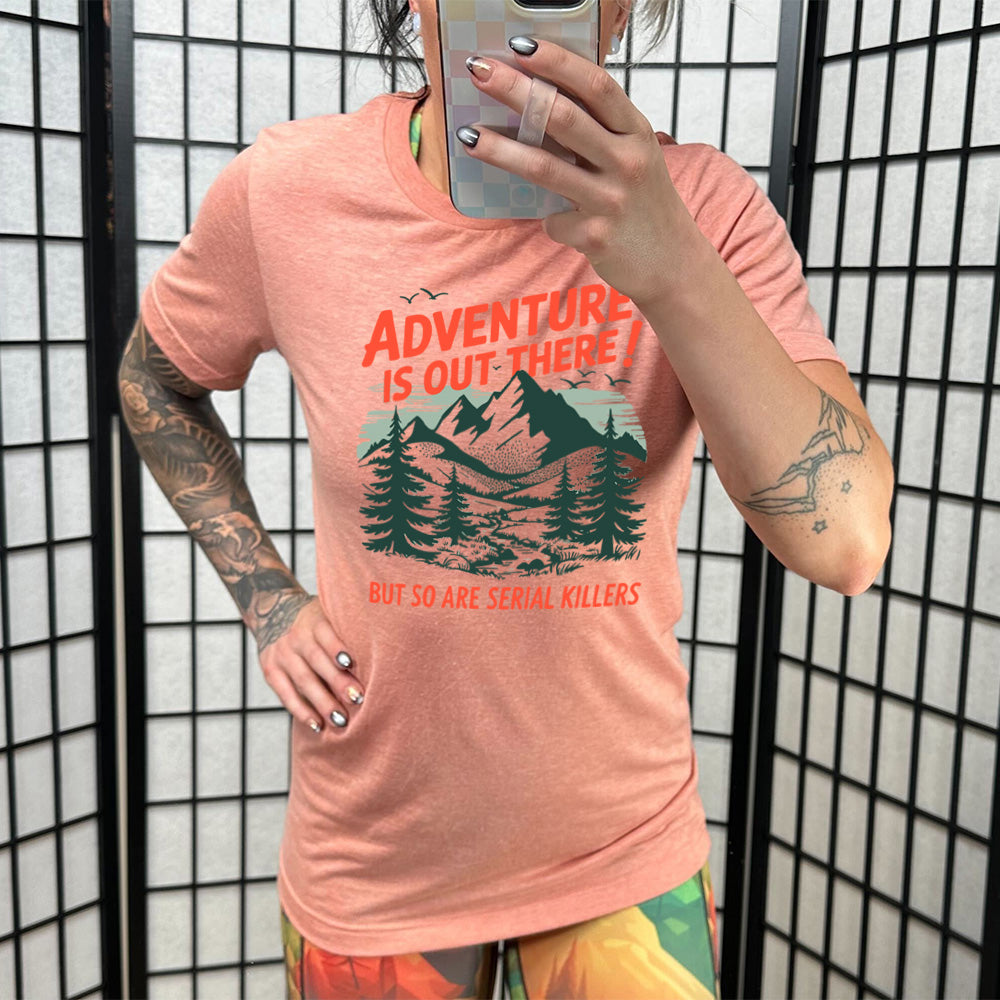 sunset unisex tee with the words "adventure is out there! but so are serial killers" on it, with the graphic of mountains and woods and birds flying 