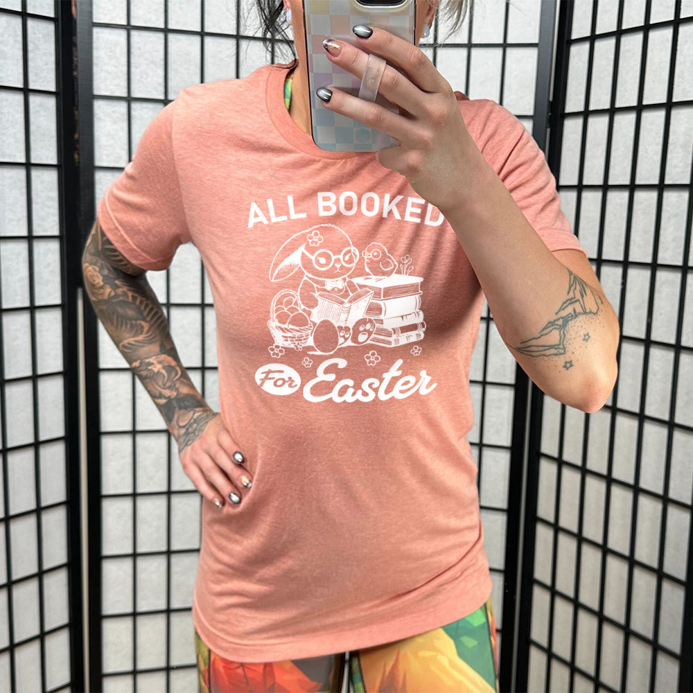sunset unisex tee with the words "all booked for Easter" on it, and the graphic of a bunny wearing glasses and a bowtie, reading a book, next to a stack of books (with a bird wearing glasses on top of it) on one side, and a basket of eggs on the other side